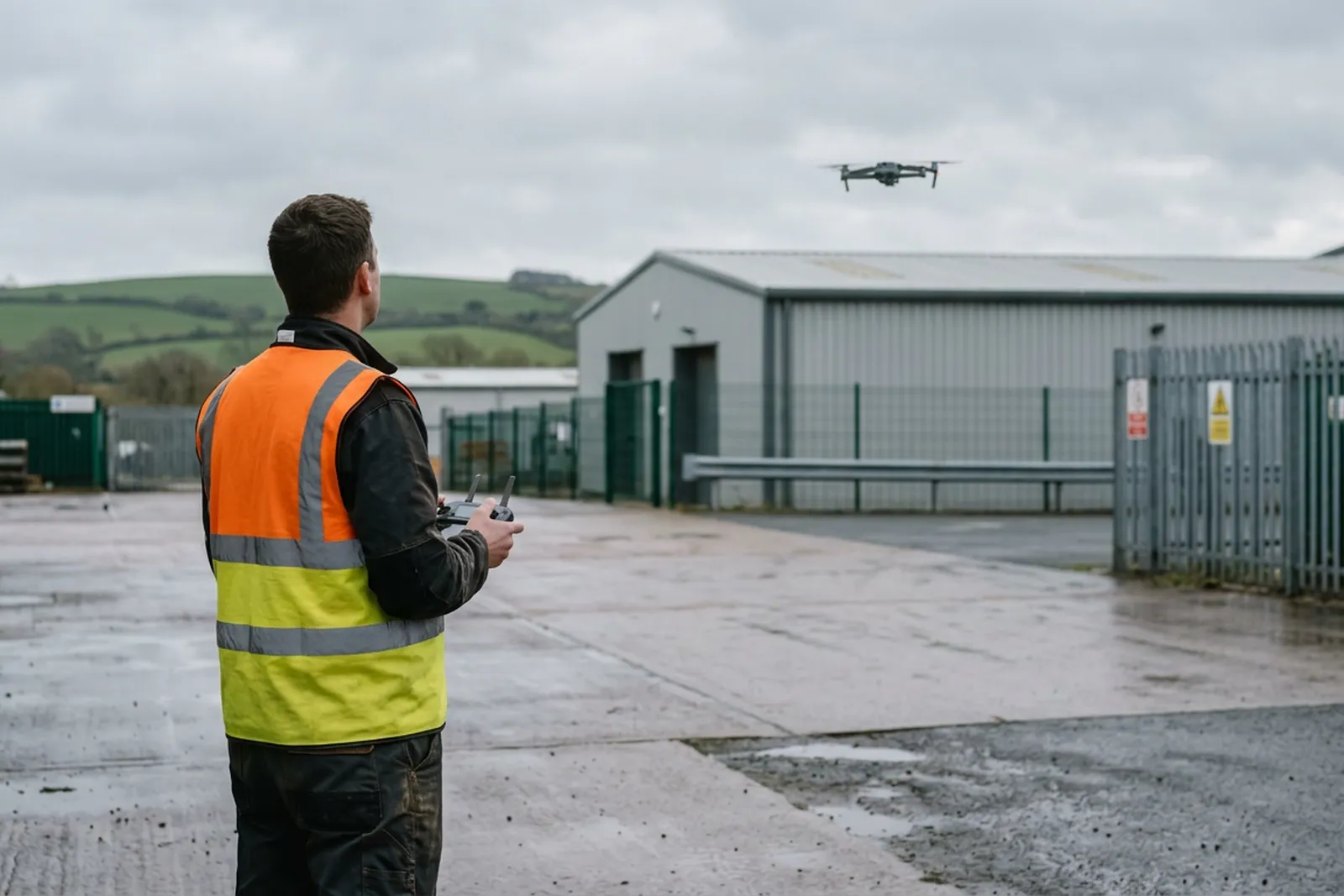 A drone pilot in a high visibility vest on a commercial yard in Devon, controller in both hands, watching a quadcopter hovering above the flat roof of an industrial unit