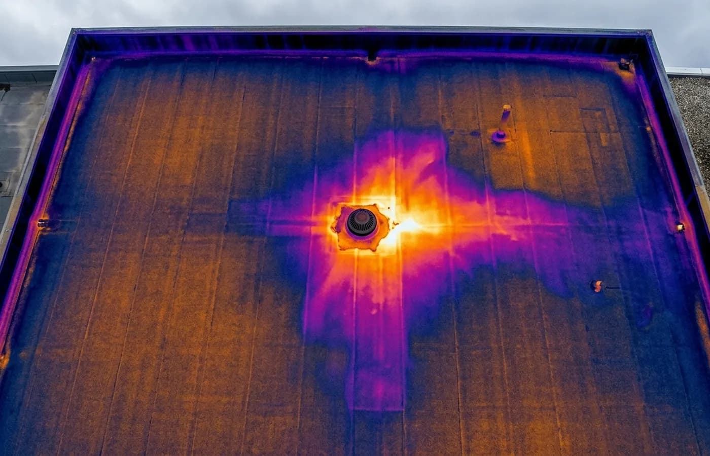 A radiometric thermal image of a flat commercial roof in ironbow false colour, showing a warm anomaly spreading beneath the membrane near an outlet